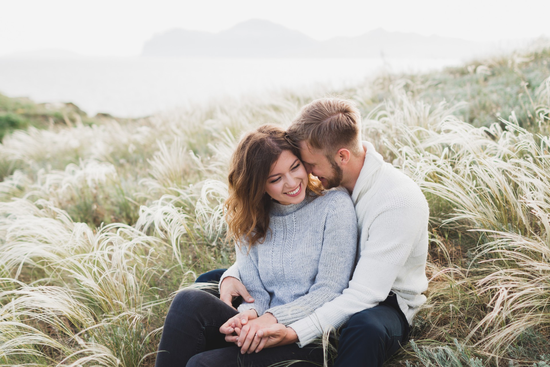 Happy young loving couple sitting in feather grass meadow, laughing and hugging, casual style sweater and jeans