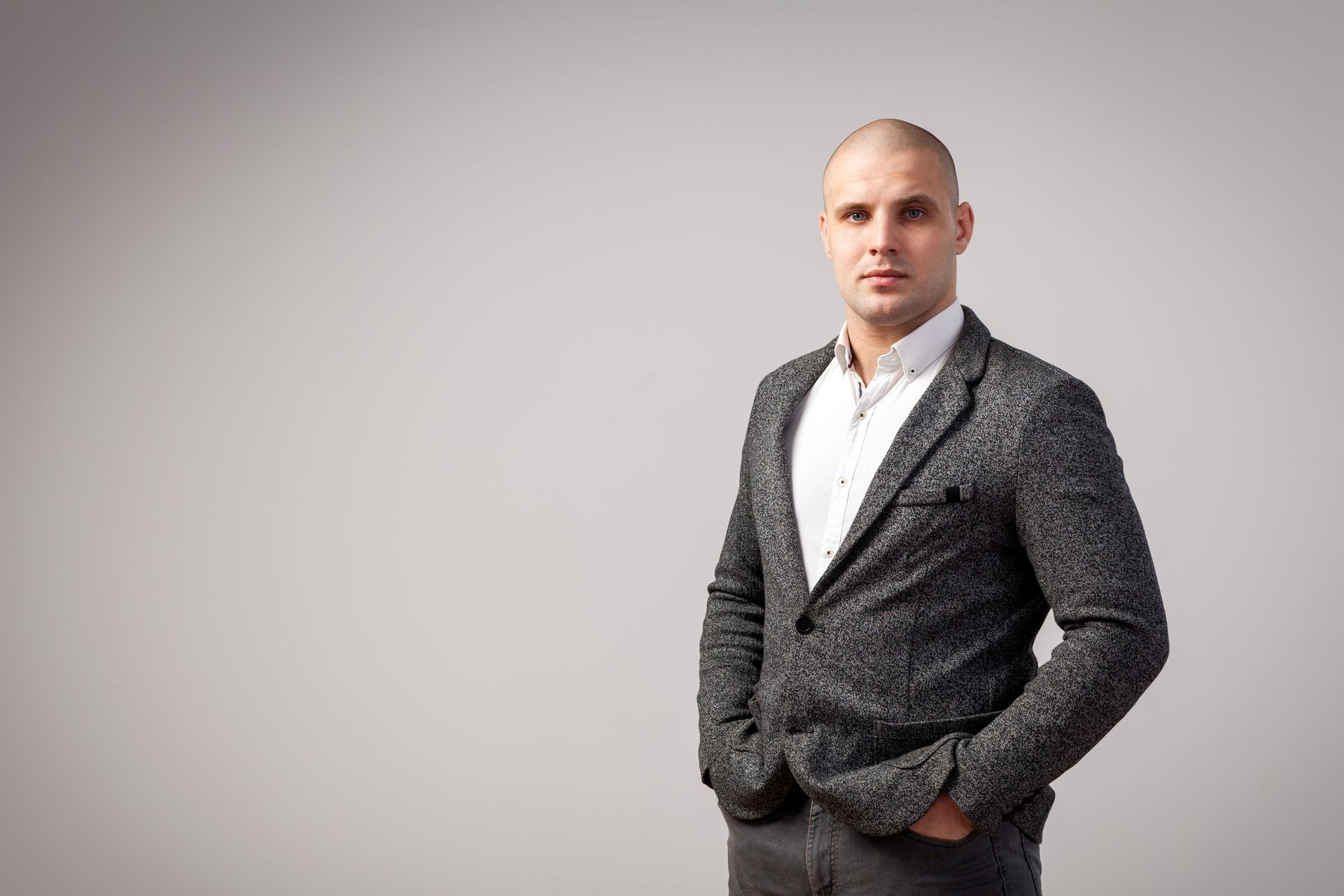 A young bald man in a white shirt, gray suit strictly looks into the camera and holds his hands in the pockets of his jacket on a white isolated background